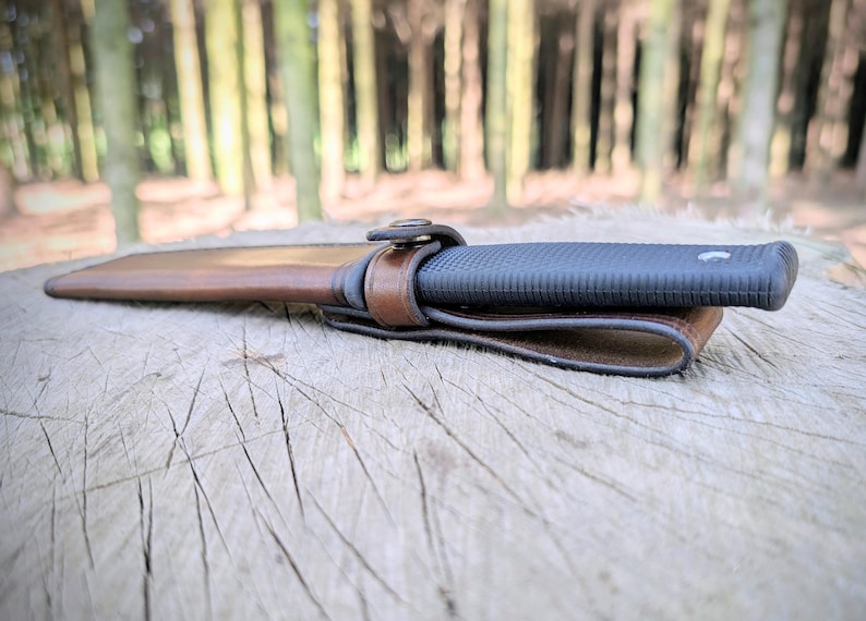 May include: A black knife with a textured handle is secured in a brown leather sheath with a matching belt loop. The knife and sheath are resting on a weathered wooden surface, with a blurred forest background.