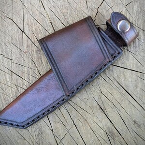 May include: A brown leather knife sheath with a snap closure. The sheath is made of a sturdy, dark brown leather and features decorative stitching along the edges. The sheath is designed to hold a knife and is shown on a wooden surface.