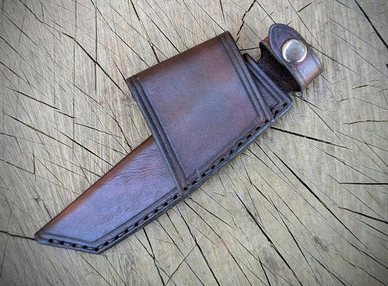 May include: A brown leather knife sheath with a snap closure. The sheath is made of a sturdy, dark brown leather and features decorative stitching along the edges. The sheath is designed to hold a knife and is shown on a wooden surface.