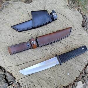 May include: A black and silver knife with a textured handle and a leather sheath. The knife has a straight blade and a black handle. The sheath is brown and has a snap closure. The knife is approximately 10 inches long.