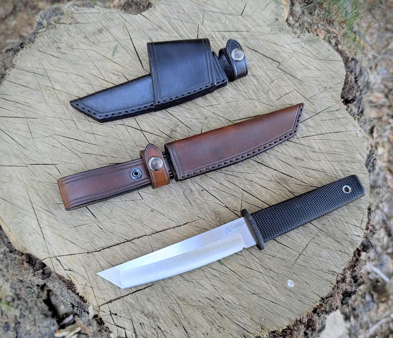 May include: A black and silver knife with a textured handle and a leather sheath. The knife has a straight blade and a black handle. The sheath is brown and has a snap closure. The knife is approximately 10 inches long.