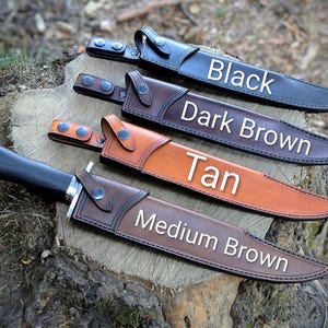 May include: Four leather sheaths in black, dark brown, tan, and medium brown, each with a knife inside. The sheaths are displayed on a tree stump. The colors are labeled with white text: Black, Dark Brown, Tan, and Medium Brown.