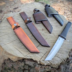 May include: A collection of knives and sheaths displayed on a tree stump. The knives have silver blades and black handles. The sheaths are made of leather in shades of brown and tan, with stitching and button closures.