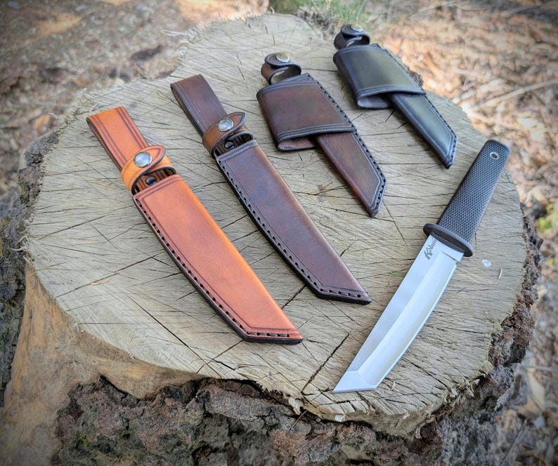 May include: A collection of knives and sheaths displayed on a tree stump. The knives have silver blades and black handles. The sheaths are made of leather in shades of brown and tan, with stitching and button closures.