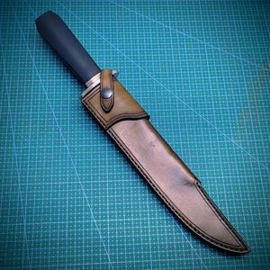May include: A handcrafted knife with a dark handle and a silver bolster, sheathed in a brown leather case. The leather sheath features a belt loop secured with a metal snap. The knife and sheath are set against a green grid background.