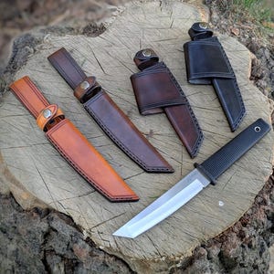 May include: An assortment of knives and sheaths displayed on a tree stump. The knives have silver blades and black handles. The sheaths are made of leather in shades of brown and black, with button closures and stitched edges.