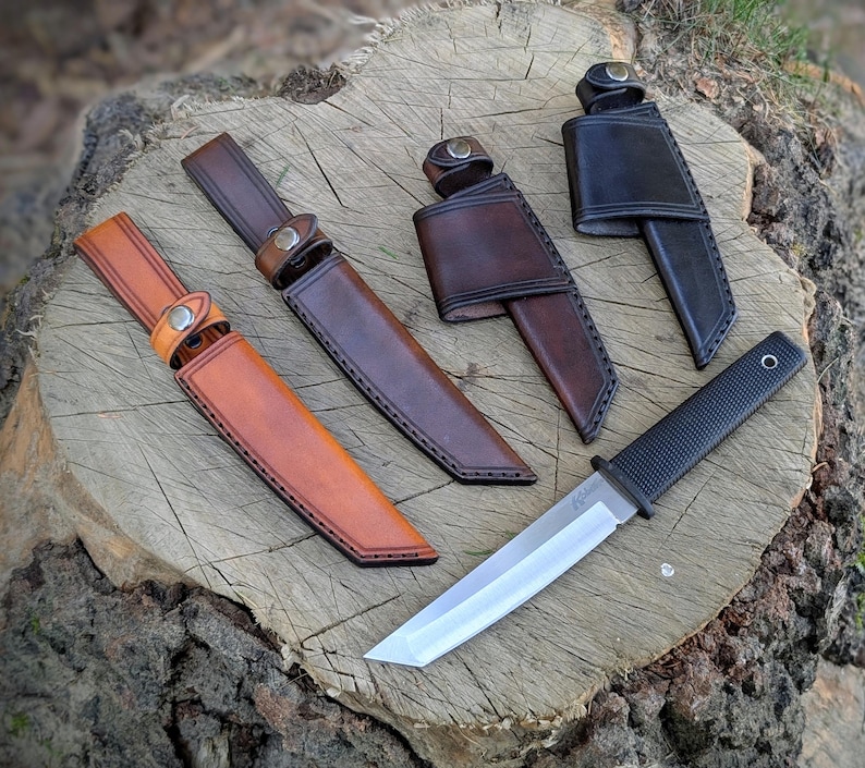 May include: An assortment of knives and sheaths displayed on a tree stump. The knives have silver blades and black handles. The sheaths are made of leather in shades of brown and black, with button closures and stitched edges.