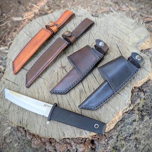 May include: A collection of knives with leather sheaths in various colors, including tan, brown, and black. The knives are displayed on a wooden surface, showcasing their craftsmanship and design. The blades are visible, and the sheaths have button closures.