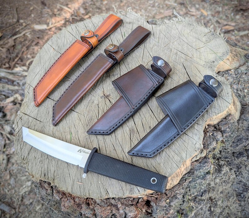 May include: A collection of knives with leather sheaths in various colors, including tan, brown, and black. The knives are displayed on a wooden surface, showcasing their craftsmanship and design. The blades are visible, and the sheaths have button closures.