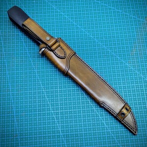 May include: A handcrafted leather knife sheath in shades of brown, with a black handle cover. The sheath features detailed stitching and metal rivets, designed to securely hold a knife. The design suggests a focus on durability and craftsmanship.