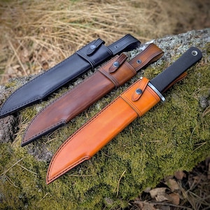 Könnte beinhalten: Drei Lederscheiden für Messer, eine schwarze, eine braune und eine orange. Die Scheiden sind alle aus Leder und haben Nähte an den Rändern.