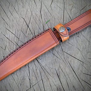 May include: A brown leather knife sheath with a matching belt loop and snap closure. The sheath has decorative stitching along the edges and is designed to hold a knife. The leather has a natural, textured appearance.