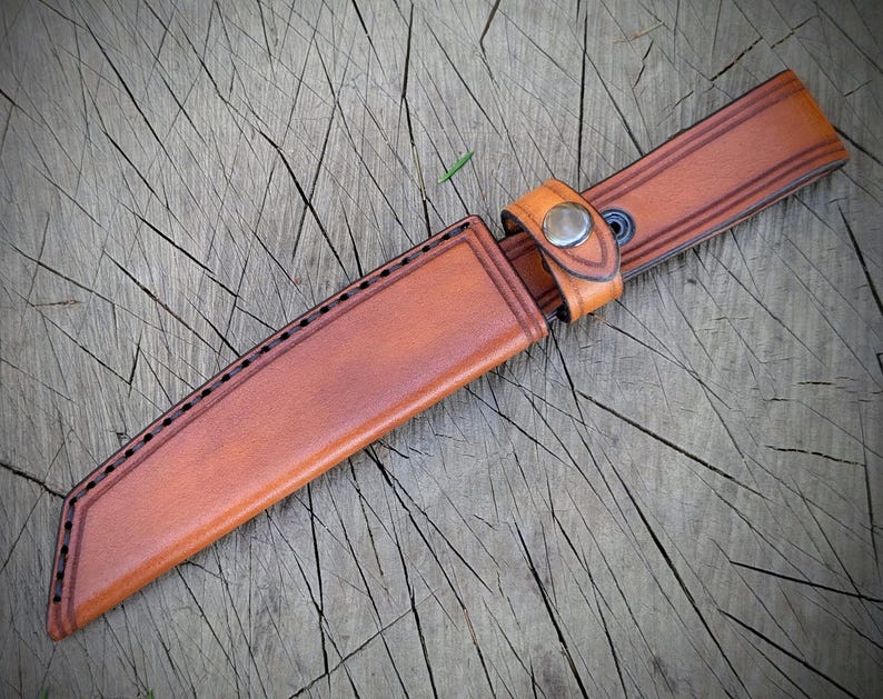 May include: A brown leather knife sheath with a matching belt loop and snap closure. The sheath has decorative stitching along the edges and is designed to hold a knife. The leather has a natural, textured appearance.