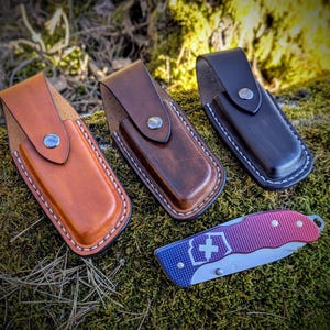 May include: Three leather knife sheaths in brown, tan, and black with white stitching. A red and blue pocket knife with a white cross on the handle is also pictured.