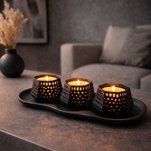 May include: Three black hexagonal candle holders with intricate cut-out patterns, each containing a lit white tea light candle. The holders are arranged in a row on a matching black oval tray, creating a warm glow through the geometric openings.