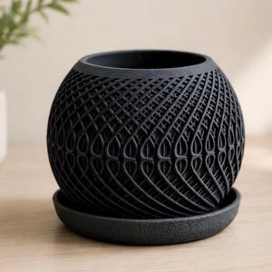 May include: A spherical black planter with a textured, intricate lattice pattern on its exterior surface. The planter sits on a matching circular black saucer with a slightly grainy finish, featuring a wide opening at the top for plant placement.