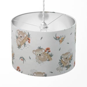 Può includere: Paralume cilindrico con un motivo ripetuto di koala dei cartoni animati, fiori e foglie su sfondo bianco. I koala sono marroni chiari e i fiori sono arancioni e gialli. Il paralume è sospeso a un cavo bianco.