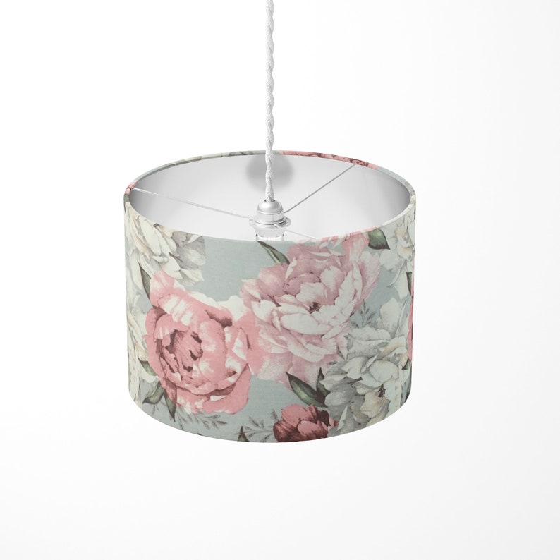 K&ouml;nnte beinhalten: Ein wei&szlig;es Kabel h&auml;ngt von einer Decke mit einer daran befestigten Leuchte. Die Leuchte hat einen runden, wei&szlig;en Stoffschirm mit einem floralen Muster aus rosa und wei&szlig;en Pfingstrosen auf hellblauem Hintergrund.
