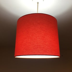 Puede incluir: Una pantalla de l&aacute;mpara de tela roja cuelga de una l&aacute;mpara de techo. La pantalla es cil&iacute;ndrica y tiene un interior blanco.