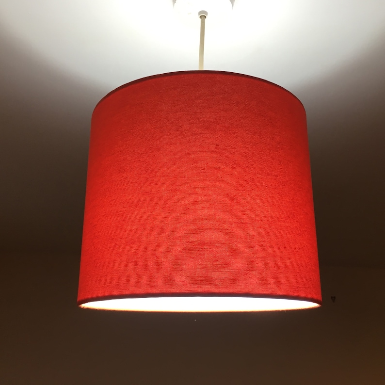 Puede incluir: Una pantalla de l&aacute;mpara de tela roja cuelga de una l&aacute;mpara de techo. La pantalla es cil&iacute;ndrica y tiene un interior blanco.