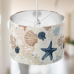 May include: A drum lampshade featuring a nautical design. The cream-coloured fabric is adorned with illustrations of seashells, starfish, and coral in shades of brown, blue, and beige. It hangs from a white cord and has a silver fitting.