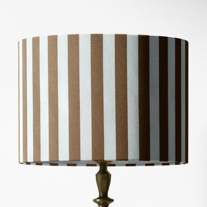 May include: A cylindrical lamp shade featuring a repeating vertical stripe pattern in alternating shades of brown and white. The shade is mounted on a metallic base with a tiered, decorative neck design, providing a structured appearance.