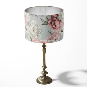 K&ouml;nnte beinhalten: Eine Tischlampe mit einem floralen Lampenschirm. Der Lampenschirm ist hellblau mit rosa und wei&szlig;en Blumen. Die Lampenbasis ist aus geb&uuml;rstetem Goldmetall.