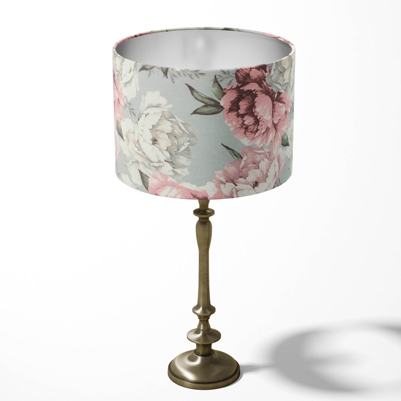 K&ouml;nnte beinhalten: Eine Tischlampe mit einem floralen Lampenschirm. Der Lampenschirm ist hellblau mit rosa und wei&szlig;en Blumen. Die Lampenbasis ist aus geb&uuml;rstetem Goldmetall.