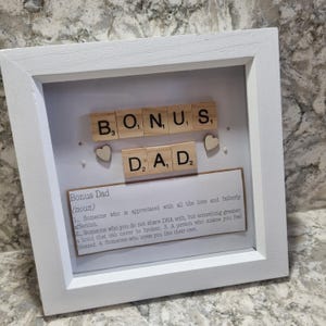 Può includere: Un'opera d'arte con cornice bianca con le parole "BONUS DAD" scritte con tessere di legno. L'opera d'arte include piccoli cuori di legno e una definizione di "Bonus Dad" su una scheda rettangolare.