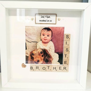 May include: A white picture frame with a photo of a baby and a dog. The frame has a small sign that says "Sandy & Ayaan, Unconditional Love XXX". The word "brother" is spelled out with wooden letter tiles.