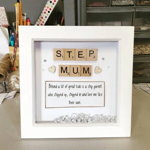 Può includere: Cornice bianca con un fronte in vetro che contiene le parole "STEP MUM" scritte con tessere di lettere in legno. La cornice include anche due piccoli cuori bianchi e una spolverata di strass trasparenti. Il testo "Behind a lot of great kids is a step parent who stepped up, stepped in and love me like their own." è stampato su uno sfondo di carta marrone.