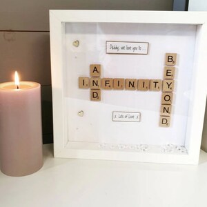 Peut inclure: Un cadre photo blanc avec un passe-partout blanc et une vitre. Le cadre contient une œuvre d'art en lettres de Scrabble qui lit "INFINITY BEYOND" avec les mots "Daddy, we love you to..." et "x. Lots of Love .x." écrits à l'encre noire. Il y a de petits cœurs argentés et des strass clairs éparpillés autour de l'œuvre d'art.