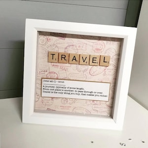 May include: A white framed print with a vintage travel theme. The word "TRAVEL" is spelled out using letter tiles. The print includes a definition of the word "travel" and a quote about travel. The background is a collage of vintage postage stamps.
