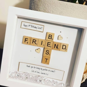 May include: A white picture frame with a white mat and a glass front. The frame contains a scrabble tile arrangement spelling out the word "FRIEND" with the word "BEST" spelled vertically. The frame also contains two small wooden hearts and a message that reads "Happy 19th Birthday Lucie!! Thank you for being my partner in crime!! Lots of Love, Lauren x x"
