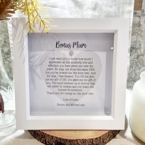May include: A white framed picture with a grey background and a heart-shaped message that reads "Bonus Mum". The message expresses appreciation for a motherly figure and the love and affection given. It includes the text "Lots of Love, Emma and Michael xxx".