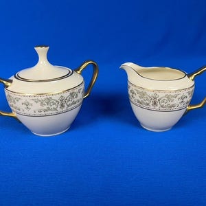 May include: A white porcelain sugar bowl with a lid and a creamer. Both pieces have gold handles and trim, and a decorative gray and gold floral band. The sugar bowl is approximately 4 inches tall.