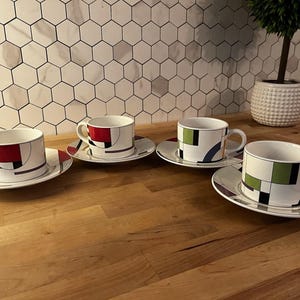 May include: Four white teacups and saucers with geometric designs in red, black, green, and blue. The cups and saucers are arranged on a wooden surface, with a white tile backsplash in the background.