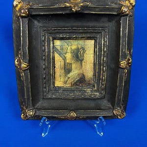 May include: A small, framed artwork featuring a profile portrait of a woman. The frame is black with gold-colored accents at the corners and edges. The artwork is displayed on a clear plastic stand against a blue background.