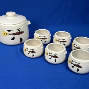May include: A ceramic set consisting of one lidded tureen and five matching small bowls. Each piece is off-white with a speckled finish and features a graphic illustration of a stylized fish in brown, black, and yellow on the exterior surface.