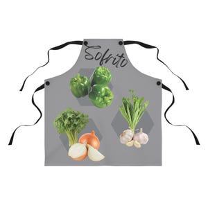 Puede incluir: Un delantal de cocina gris con tirantes negros y la palabra "Sofrito" impresa en negro. El delantal presenta imágenes de pimientos verdes, cilantro, cebollas y ajos.
