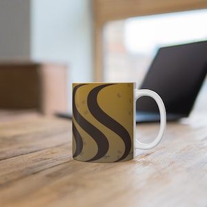May include: White ceramic coffee mug with a brown and gold design. The design features curved, wavy lines and small circular shapes. The mug is sitting on a wooden surface, with a laptop in the background.