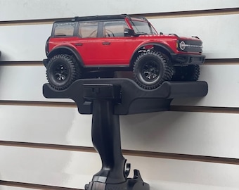 May include: A red remote control car with black wheels and a black remote control. The car is on a black wall mount.