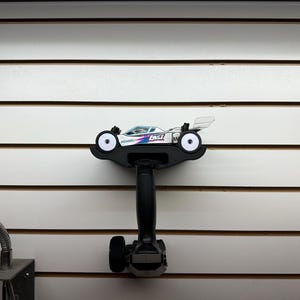 May include: A white and blue RC car with the word "LOSI" on the side is mounted on a black remote control. The remote control is mounted on a white wall with horizontal lines. The RC car has a clear wing.