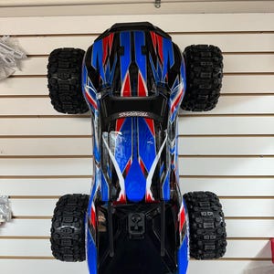 May include: A blue, red, and white remote control monster truck with black tires. The truck is hanging from the ceiling.
