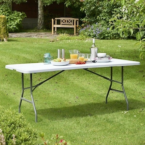 May include: White folding table with black metal legs set up outdoors on a grassy lawn. The table is set with dishes, glasses, and food.