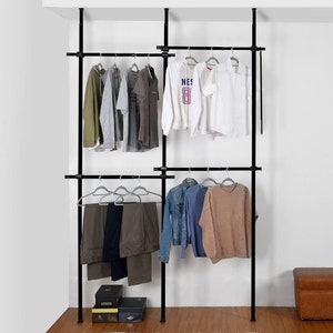 May include: A black metal clothing rack with two adjustable shelves. The rack is filled with various clothing items, including shirts, trousers, and a jumper. The rack is standing on a wooden floor.