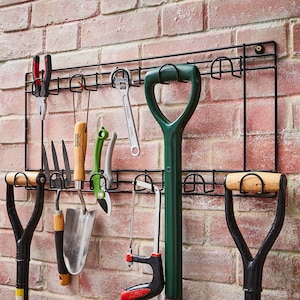 May include: A black metal wall-mounted rack with hooks for hanging gardening tools. The rack has a variety of tools hanging on it, including a green trowel, a pair of pruning shears, a hand rake, and a small hand shovel.