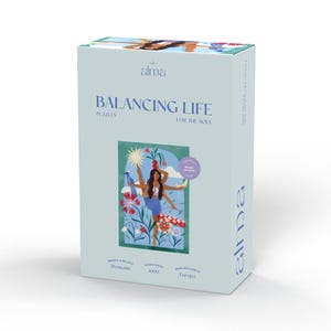Könnte beinhalten: Eine blaue Box mit einer Frau, die auf einer Hand balanciert, umgeben von einer Sonne, einem Mond und Blumen. Die Box trägt die Aufschrift "Balancing Life Puzzles for the Soul" und "Moments 1000 Puzzle Pieces Made with Love in Europe".