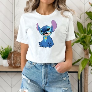 May include: A white t-shirt with a blue cartoon character wearing a yellow collar. The character has large blue eyes, a purple nose, and a big smile. The character is sitting with its legs crossed and its arms crossed.