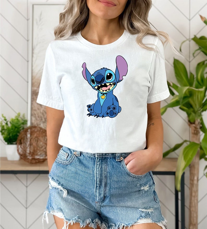 May include: A white t-shirt with a blue cartoon character wearing a yellow collar. The character has large blue eyes, a purple nose, and a big smile. The character is sitting with its legs crossed and its arms crossed.